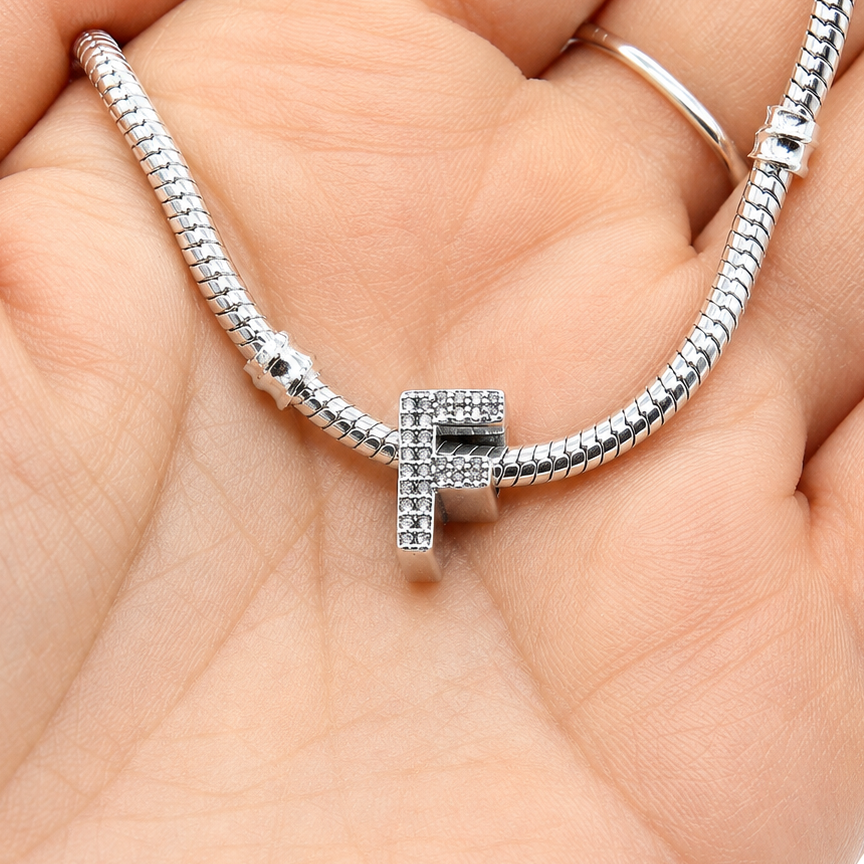 A silver snake chain bracelet with a central letter 'F' charm paved with small clear stones, resting on a person's palm.