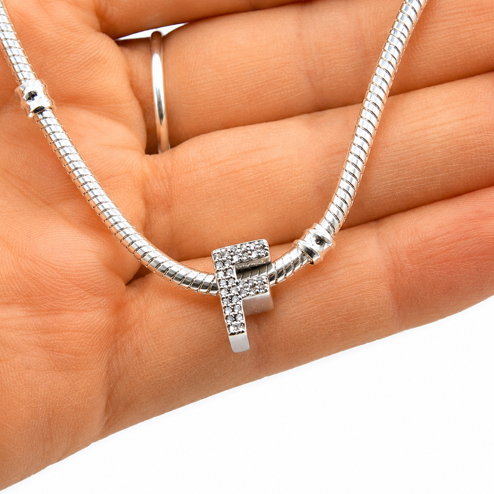 A silver snake chain bracelet with a small sterling silver letter F-shaped charm covered in clear rhinestones, held in a person's hand against a white background.