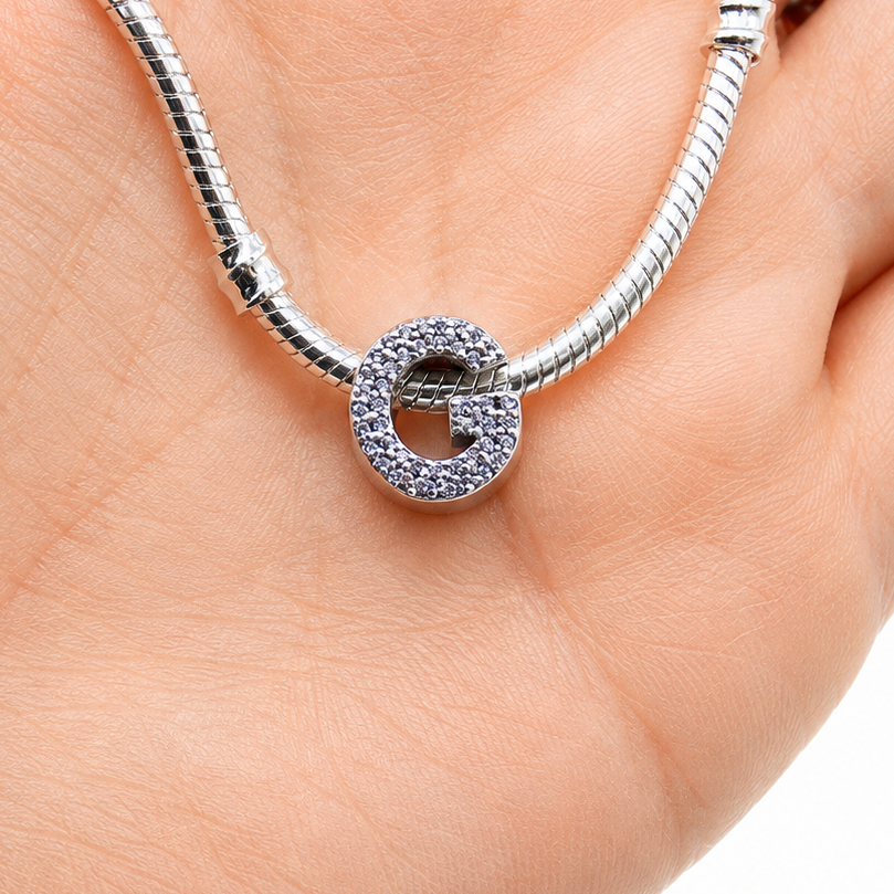 A silver snake chain necklace with a sterling silver letter 'G' initial charm paved with small clear stones, held in the palm of a hand against a white background.