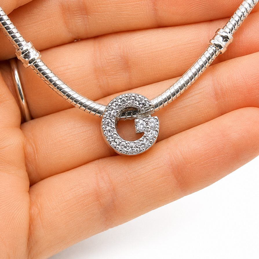 A silver snake chain necklace with a sterling silver pavé-set letter G charm, held in a person's hand against a white background.