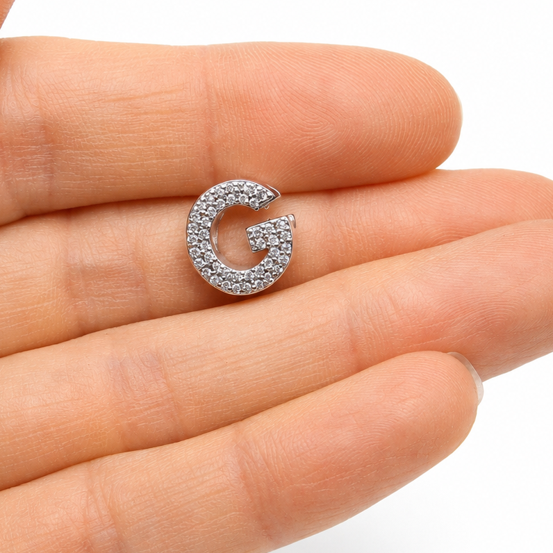 Sterling silver letter 'G' initial charm with cubic zirconia stones covering the front of the letter, held on the tip of a person's finger against a white background.