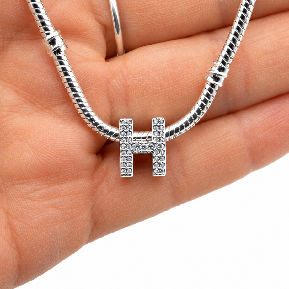 A silver snake chain bracelet with a sterling silver letter H initial charm set with small clear stones, held in a person's hand against a white background.