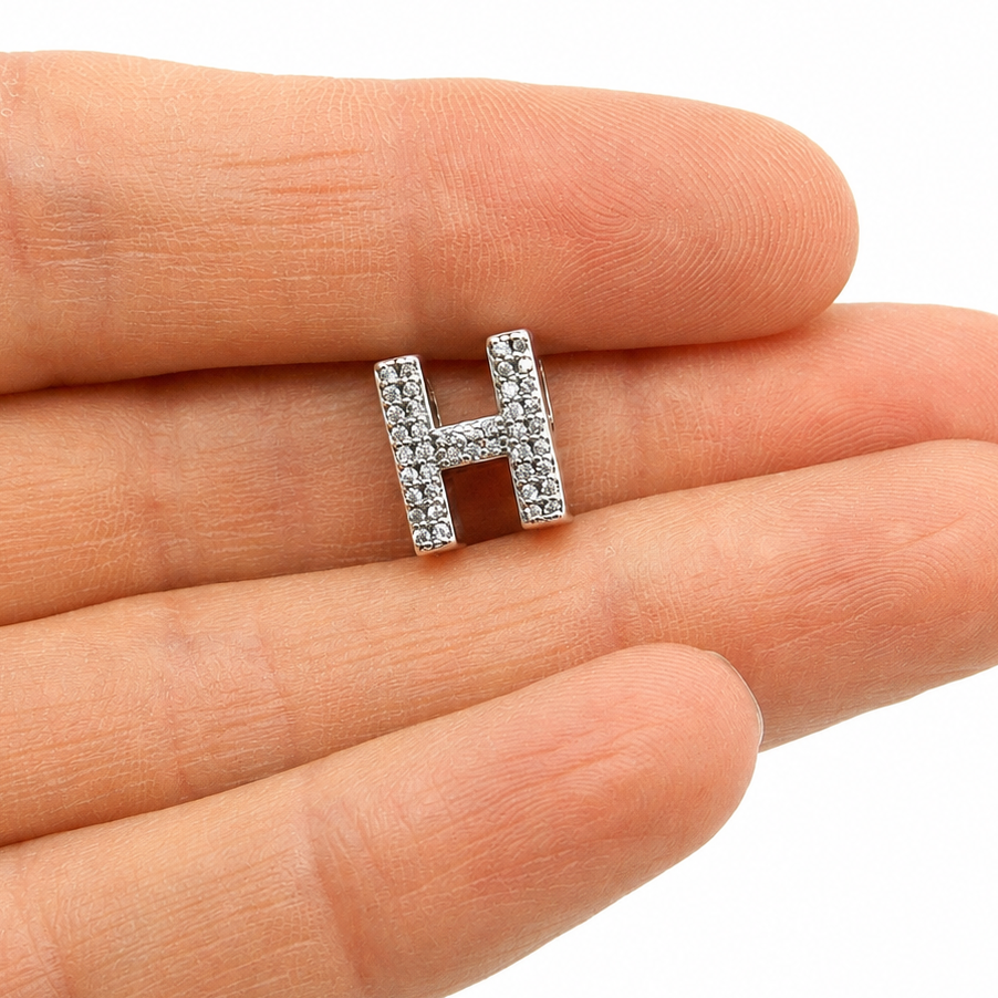 A sterling silver letter H initial charm paved with small clear stones, held on the fingertips of a person's hand against a white background.