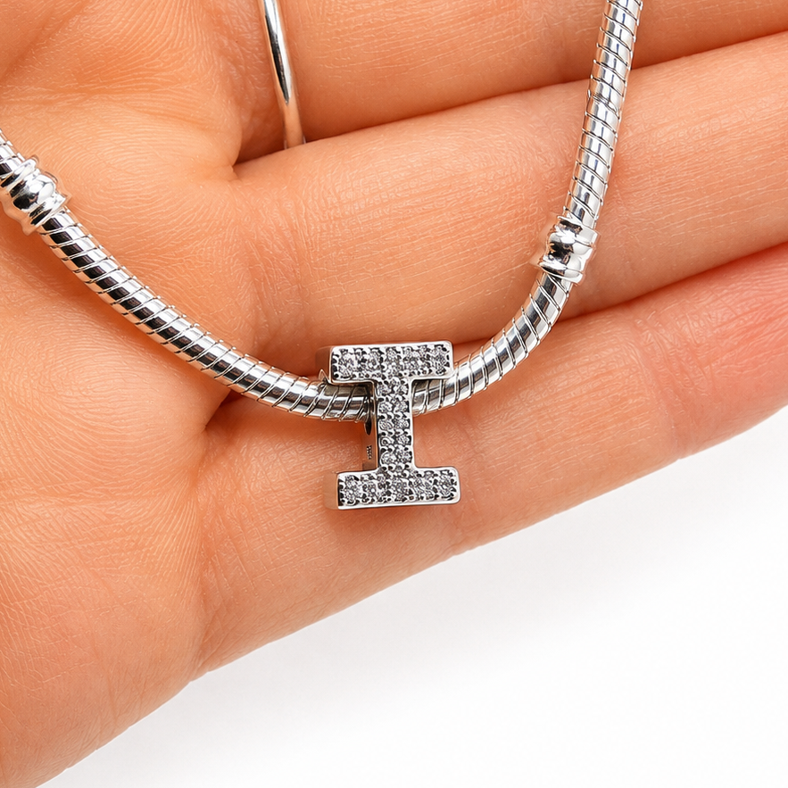 A silver snake chain bracelet with a sterling silver letter 'I' initial charm paved with small clear stones, held in the palm of a hand on a plain white background.