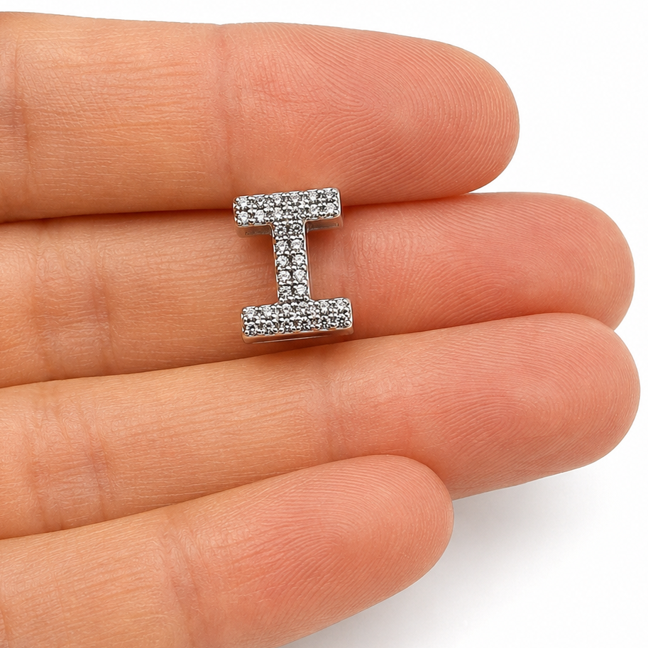 A sterling silver letter 'I' initial charm paved with clear stones, shown resting on a person's fingers against a white background.