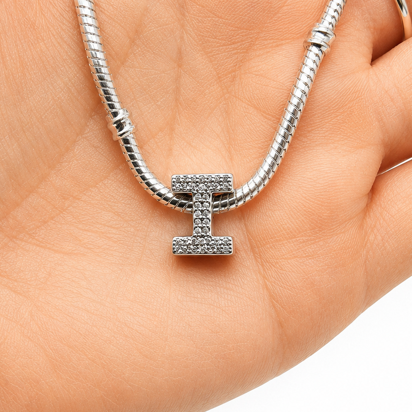 A silver-toned snake chain bracelet with a block-letter 'H' charm, displayed on a person's palm. The charm is embellished with small clear stones.
