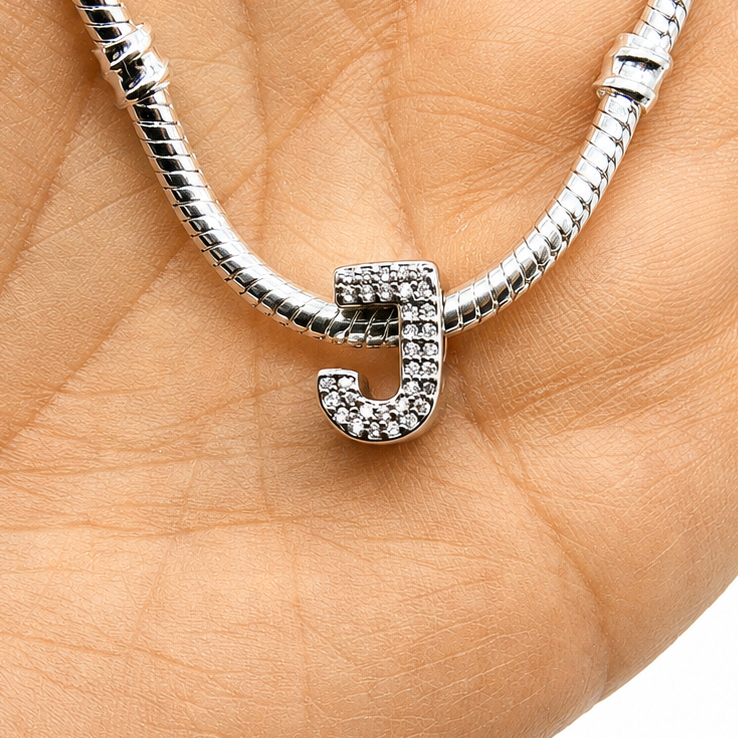 A silver snake chain bracelet with a small, sterling silver rhinestone-encrusted letter 'J' charm, held in the palm of a hand against a white background.