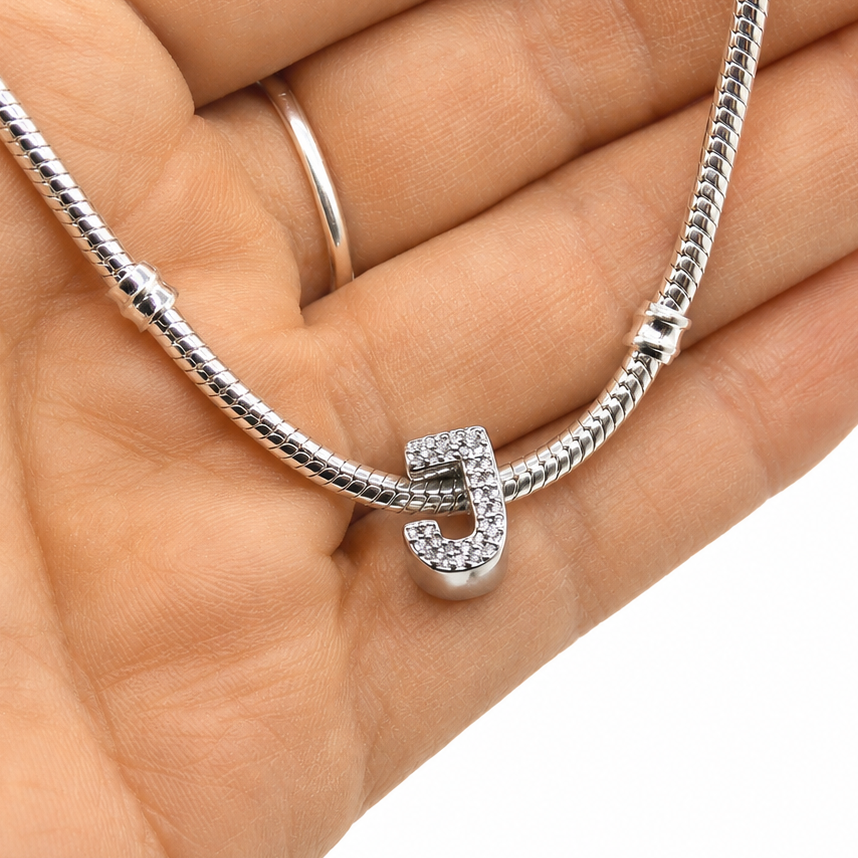 A silver snake chain bracelet with a small sterling silver letter 'J' charm paved with clear rhinestones, held in a person's hand against a white background.