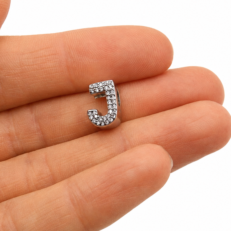 A sterling silver letter 'J' initial charm encrusted with small clear stones, held on the tip of a person's finger against a white background.