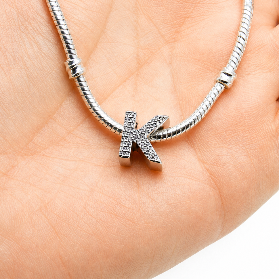 A silver snake chain bracelet with a dangling letter 'K' charm paved with small clear stones, shown resting on a person's palm.