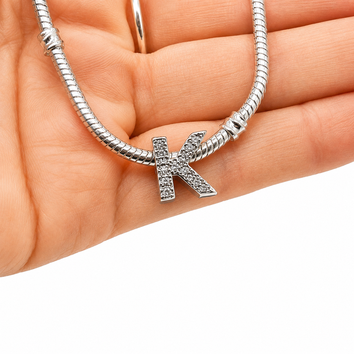 A silver snake chain necklace with a sterling silver letter 'K' initial charm paved with small clear stones, held in a person's hand against a white background.