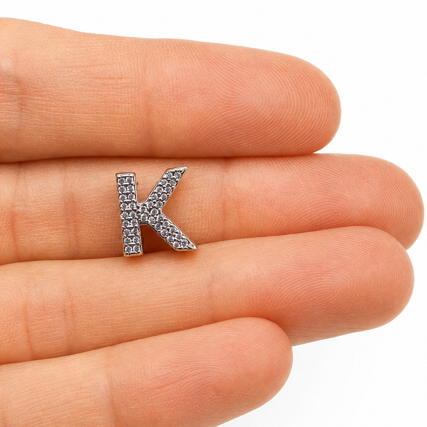 A silver letter K initial charm paved with small clear stones, held on the fingertips of an open hand to show scale.
