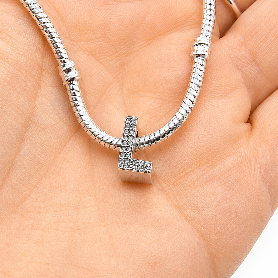 A silver snake chain bracelet with a letter 'L' charm paved with small clear stones, resting on a person's palm.
