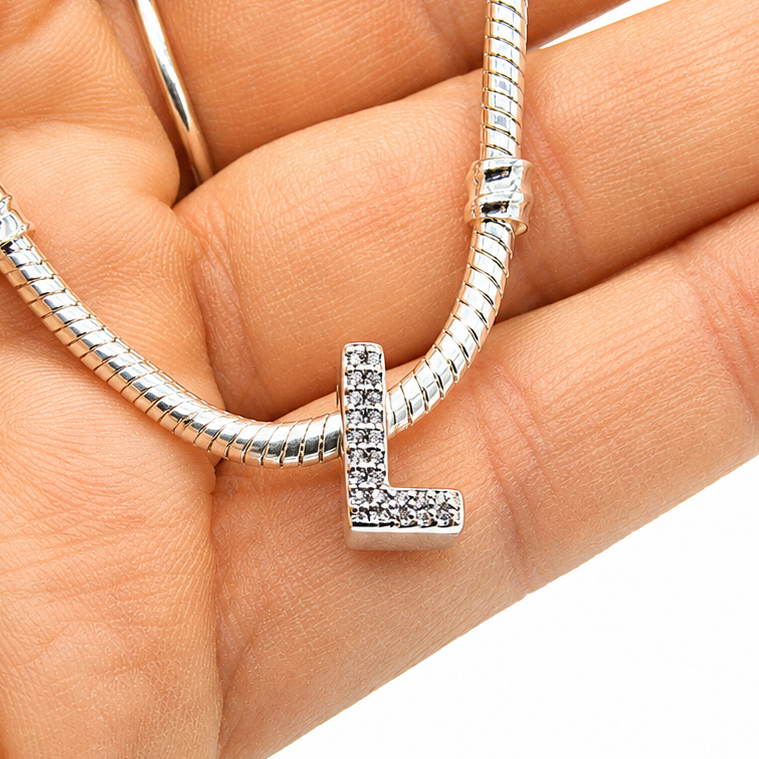 A silver snake chain necklace with a letter 'L' initial charm paved with small clear stones, held in a person's hand against a white background.
