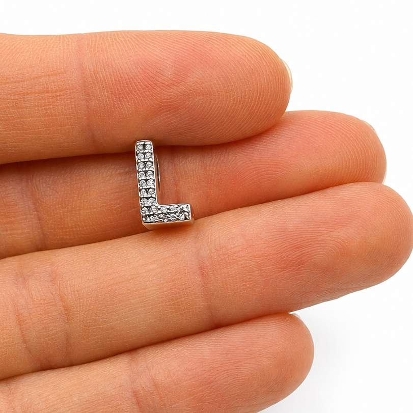 A small sterling silver letter 'L' initial charm paved with clear stones, resting on the fingertips of a hand for scale against a white background.
