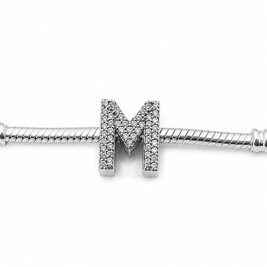 A sterling silver initial charm featuring a letter 'M' paved with small clear stones, shown on a silver snake chain against a white background.