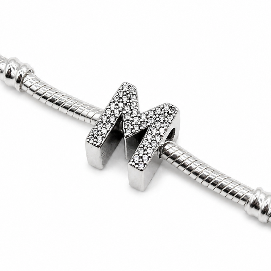 A sterling silver initial charm bead featuring the letter 'M' encrusted with small clear stones, threaded onto a silver snake chain bracelet against a white background.
