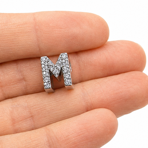 A sterling silver letter 'M' initial charm embellished with pavé-set clear stones, one person's fingertips for scale, shown on a clean white background.