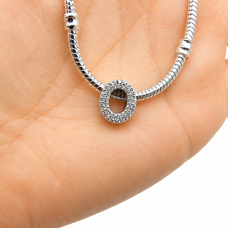 A silver snake chain necklace with a sterling silver letter 'O' charm bead embellished with clear rhinestones displayed on a person's open palm against a white background.